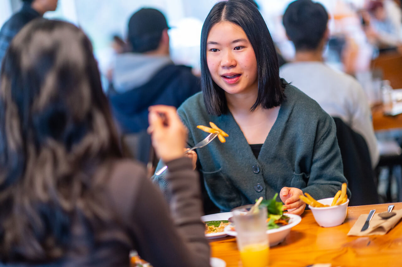 A young woman facing us, eating fries and salad while speaking to a friend who is facing away from us.