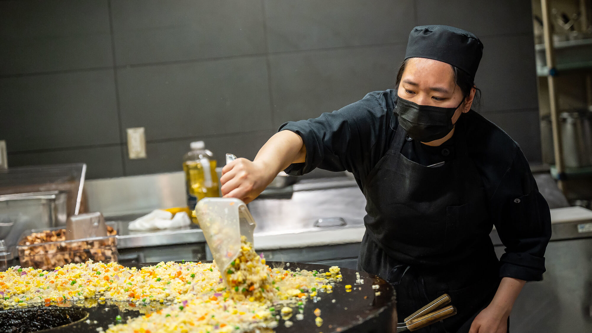 Photograph of a feminine-presenting person with a medium skin tone, wearing all-black chef's clothes and apron. Her long black hair is tied back and she wears a short, black, pillbox-style chef hat. She is cooking a batch of fried rice on a large Mongolian-style grill at Open Kitchen dining hall in Orchard Commons residence at UBC Vancouver.